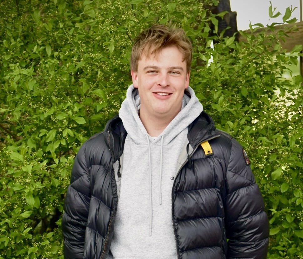 Eine Person in schwarzer Daunenjacke lächelt vor einem Hintergrund aus üppigem, grünem Laub.