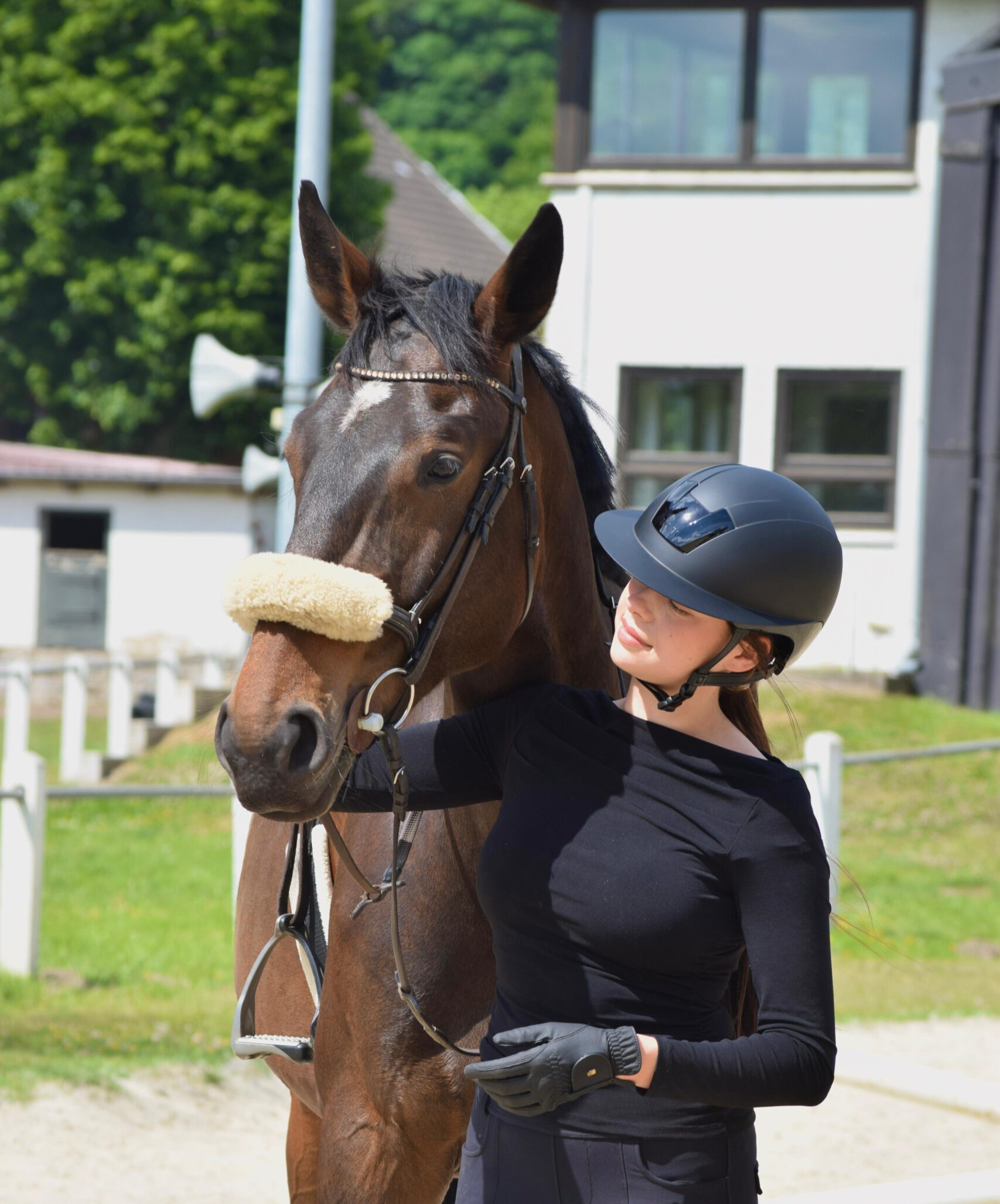Eine Frau in Reitkleidung steht neben einem braunen Pferd und berührt es sanft in ländlicher Umgebung.