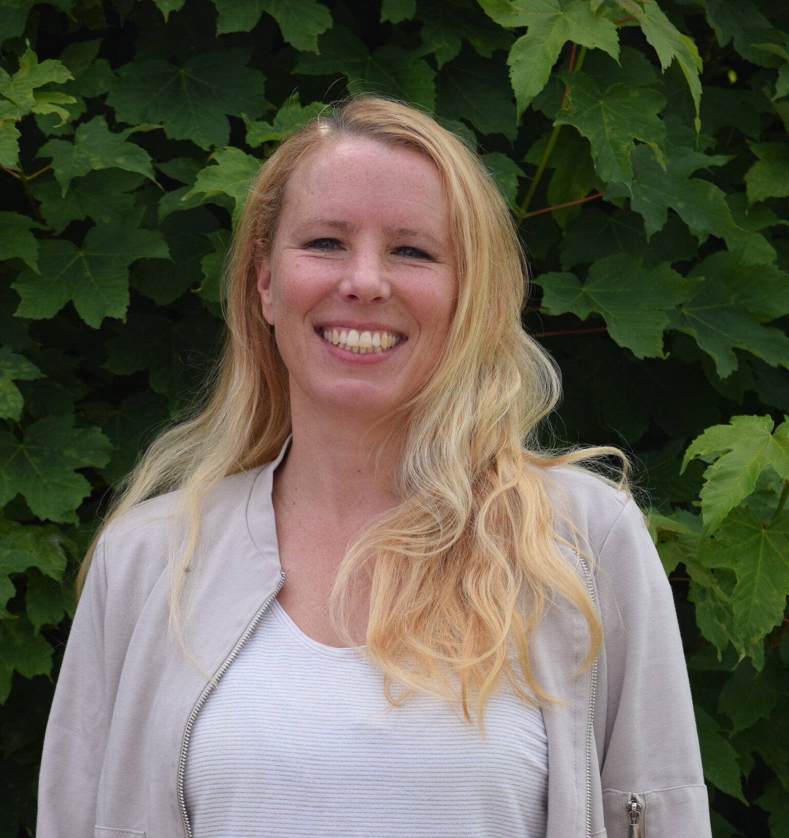 Eine lächelnde Person mit blonden Haaren steht vor einem Hintergrund aus großen, grünen Blättern.