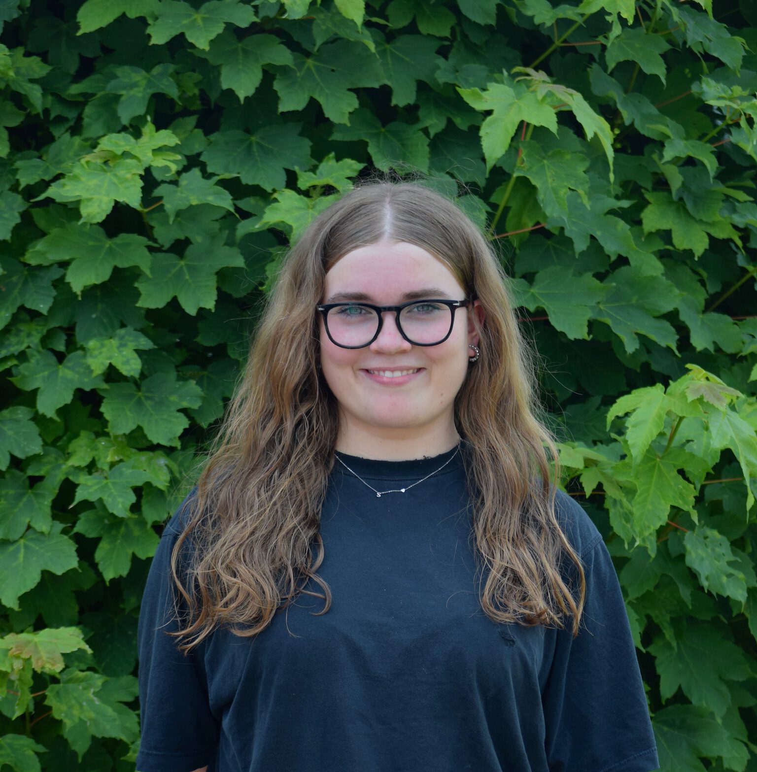 Person mit Brille lächelt vor grüner Laubwand, trägt schwarzes T-Shirt und hellblaue Jeans.