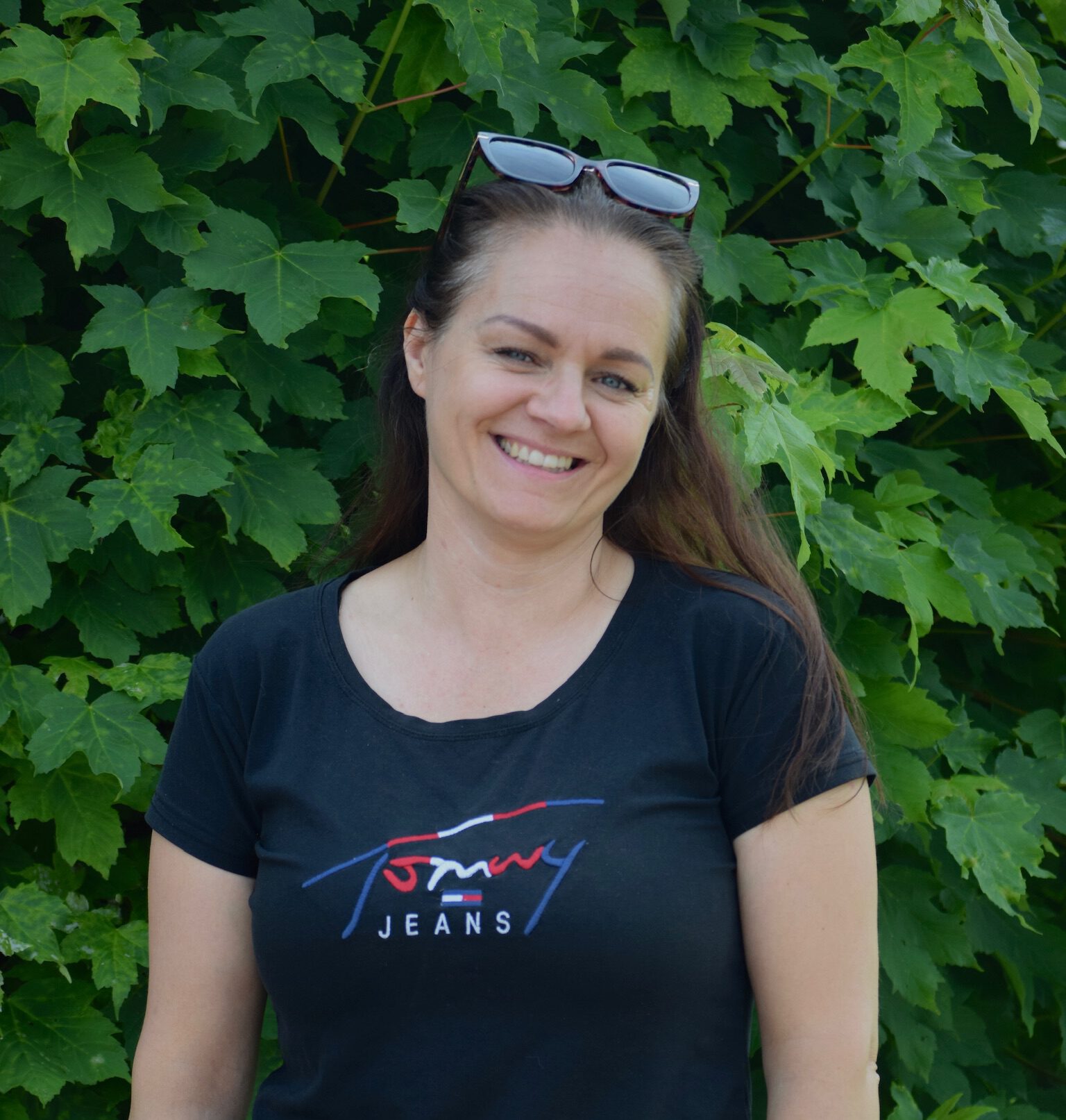 Frau mit schwarzem "Tommy Jeans"-T-Shirt lächelt vor grüner Blätterwand in die Kamera.