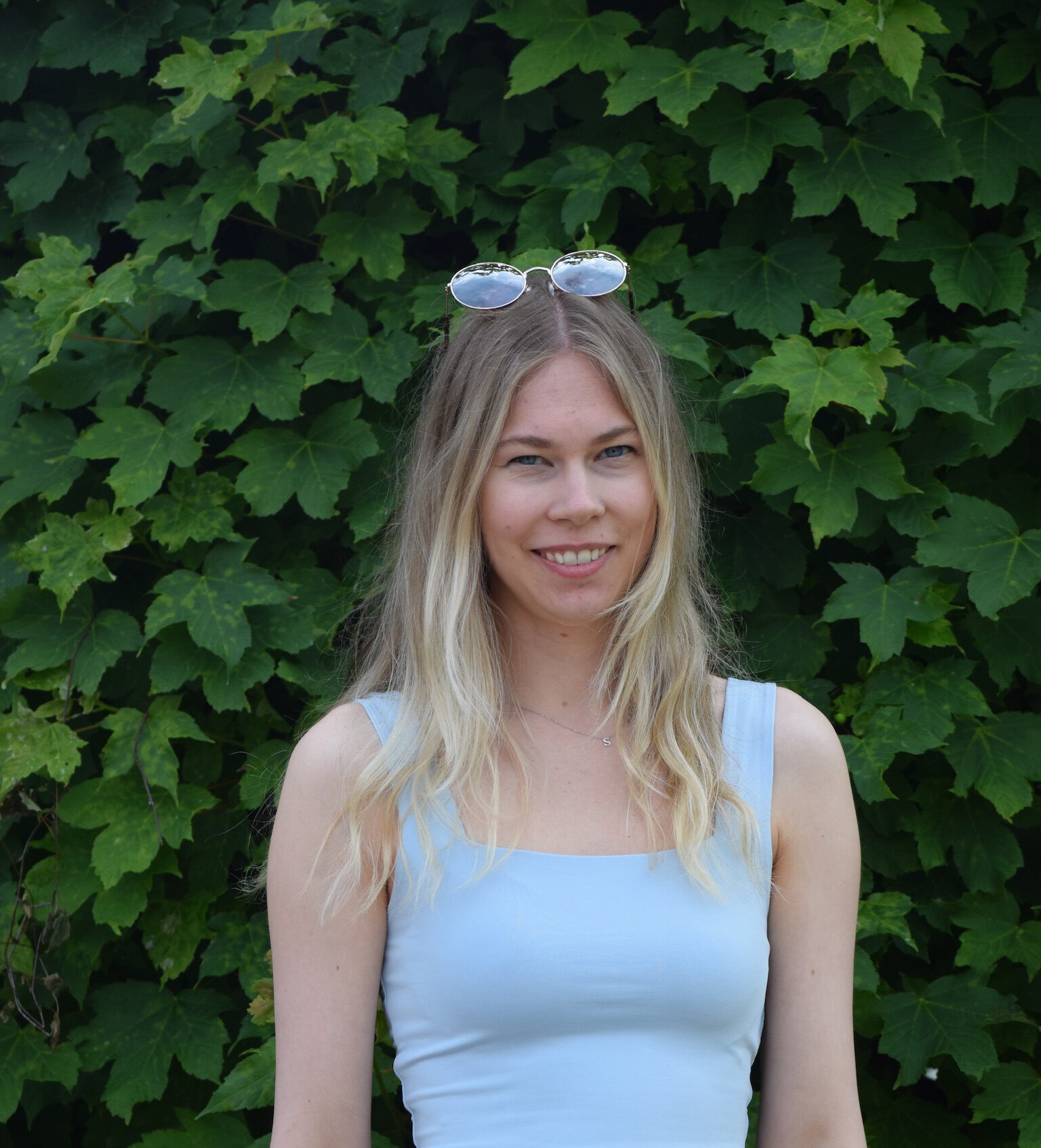 Eine lächelnde Person mit blondem Haar steht vor grünem Laub, trägt ein blaues Top und eine Sonnenbrille.
