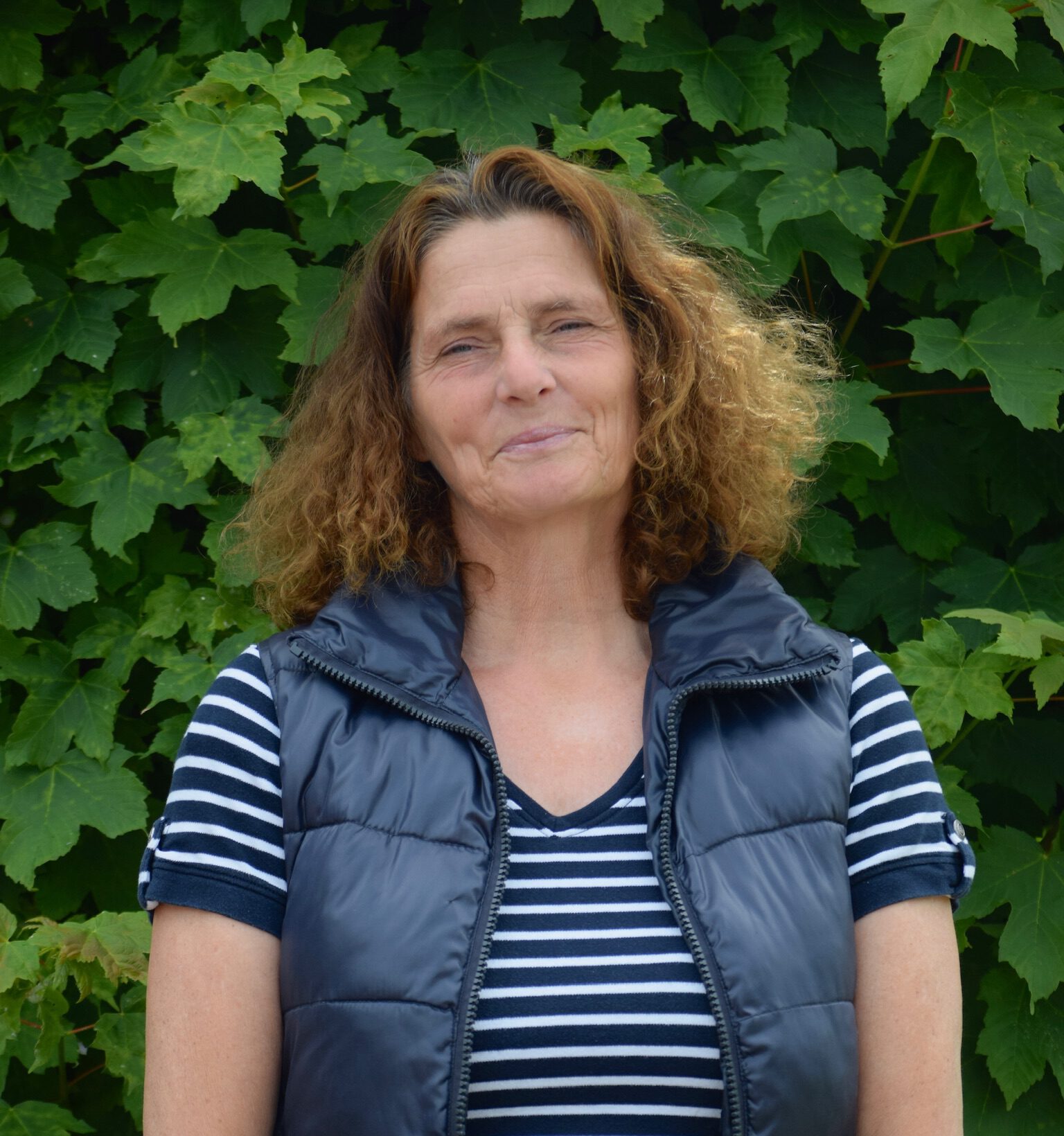 Eine Frau mit lockigem Haar lächelt leicht vor einem Hintergrund aus grünen Blättern.